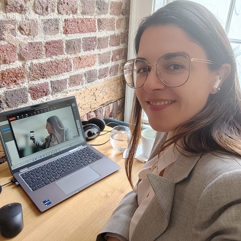 Stefania Arca smiling while working remotely at a café with a laptop and brick wall backdrop, showcasing a flexible and modern work style
