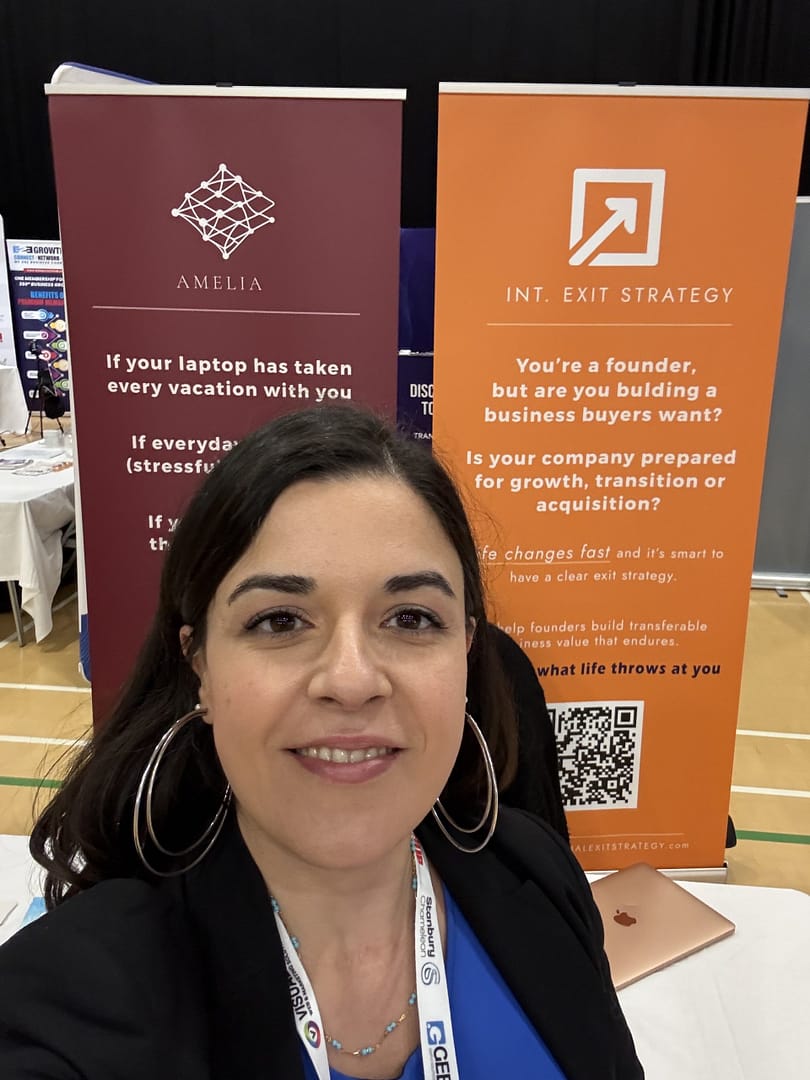 Close-up portrait of Stefania Arca, founder of International Exit Strategy, at a professional business exhibition in the UK, showcasing branded company banners about exit strategy consulting and business systemisation.